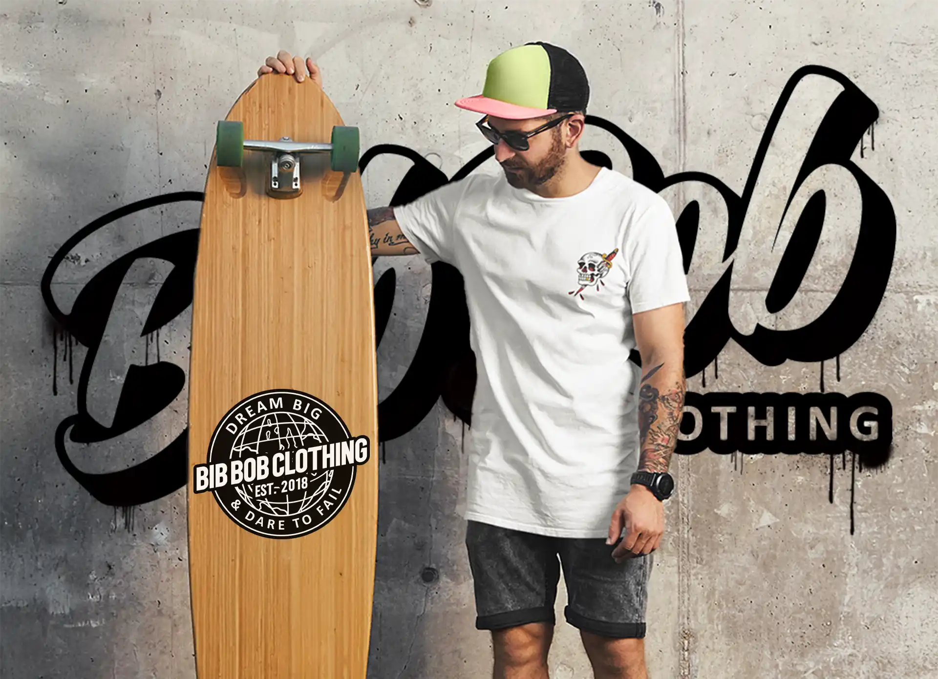 A man wearing a white t-shirt, dark shorts, and a two-tone baseball cap, holding a wooden longboard with green wheels.